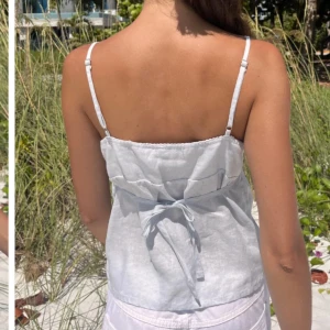 Vit linnetopp med knytband brandy mellvie - Säljer en stilren vit linnetopp med smala axelband och knytband i ryggen. Toppen har en v-ringad design och är perfekt för varma sommardagar.