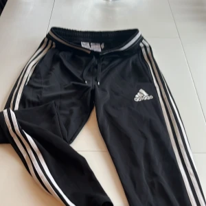 Svarta träningsbyxor från Adidas - Snygga svarta träningsbyxor från Adidas med de klassiska tre ränderna längs sidorna. Byxorna har en elastisk midja med snörning och är perfekta för en sportig look.
