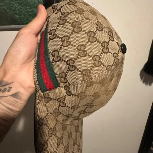 Beige keps med monogrammönster - Snygg beige keps från Gucci med det ikoniska monogrammönstret. Kepsen har en justerbar rem i skinn baktill och en grön och röd randdetalj på sidan. Perfekt för att ge din outfit en lyxig touch.