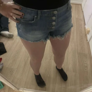 Blå jeansshorts med knappar - Snygga blå jeansshorts med hög midja och fransiga kanter. De har en knappgylf med flera knappar framtill och klassiska femficksdetaljer. Perfekta för en avslappnad sommarlook.