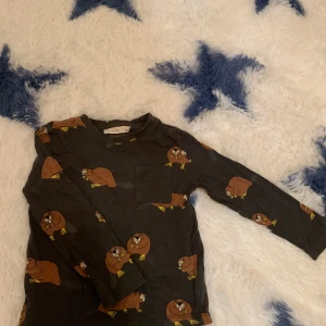 Grå långärmad tröja med bävermönster från Mini Rodini - Säljer en grå/brun långärmad tröja från Mini Rodini med ett roligt bävermönster. Perfekt för barn som gillar djurmotiv. Tröjan har en liten bröstficka och är gjord i mjukt material för extra komfort.