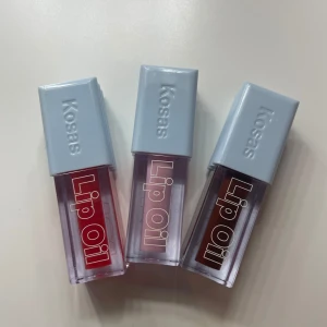 Kosas Wet Lip Oil 3st - Säljer tre olika nyanser av Kosas Lip Oil - Cola, Sorbet och Slushy. Förpackningarna är stilrena med ljusgrå lock och transparenta behållare som visar färgerna: en djup röd, en mjuk rosa och en mörkare brun. Perfekt för att ge läpparna en glansig och vårdande finish. 