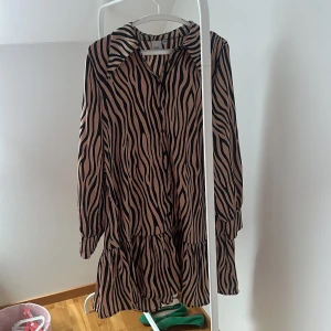 Zebramönstrad klänning från ICHI - Säljer en trendig zebramönstrad klänning från ICHI. Klänningen har långa ärmar och en volangdetalj vid nederkanten. Perfekt för att sticka ut med sitt djärva mönster och bekväma passform.