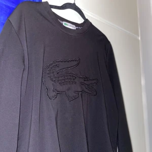 Svart tröja från Lacoste - Säljer en svart tröja från Lacoste med det ikoniska krokodilmärket i svart på bröstet. Tröjan har långa ärmar och en rund halsringning. Perfekt för en stilren look. Passar även storlek S