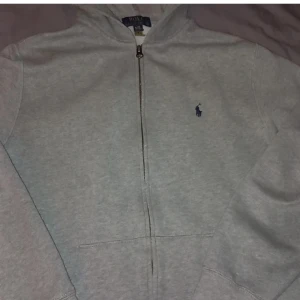 Grå hoodie från Polo Ralph Lauren - Säljer en klassisk grå hoodie från Polo Ralph Lauren med dragkedja och broderad logga på bröstet. Perfekt för en avslappnad stil. Hoodien har långa ärmar och en bekväm passform.