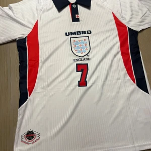 Vit fotbollströja från Umbro - Säljer en vit fotbollströja från Umbro med England-emblem och nummer 7 på framsidan. Tröjan har korta ärmar och röda detaljer längs sidorna. Perfekt för fotbollsfans!