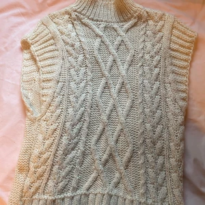 Beige stickad väst - Säljer en mysig beige stickad väst med ett snyggt flätmönster på framsidan. Västen har en hög krage och ribbade kanter vid ärmhålen. Perfekt för lager-på-lager under kyligare dagar.