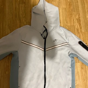 Ljusgrå hoodie från Nike - Säljer en ljusgrå hoodie från Nike med dragkedja och huva. Jackan har en svart randdetalj över bröstet och en ficka på ärmen. Perfekt för en avslappnad stil.