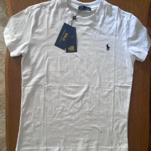 Vit t-shirt från Ralph Lauren - Säljer en klassisk vit t-shirt från Ralph Lauren med den ikoniska broderade loggan på bröstet. T-shirten är kortärmad och tillverkad i mjuk bomull. Perfekt för en stilren och avslappnad look.
