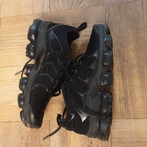 Svarta air vapormax plus - Säljer ett par svarta Nike sneakers med unik sula och snörning. Skorna har en modern design med synliga luftkuddar i sulan för extra komfort. Perfekta för en sportig look.