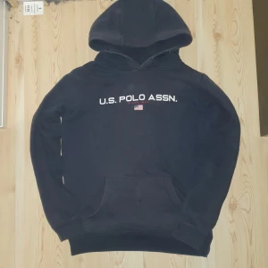 Mörkblå hoodie från U.S. Polo Assn. - Säljer en mörkblå hoodie från U.S. Polo Assn. med vit text och en liten amerikansk flagga på bröstet. Tröjan är tillverkad i en mjuk bomullsblandning och har en klassisk känguruficka framtill. Perfekt för en avslappnad stil.
