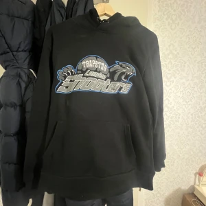  Trapstar Shooterz Tracksuit – Svart & blåa  - Ge din outfit en streetwear-edge med denna Trapstar Shooterz Tracksuit i svart och blått. Designad för både stil och komfort, med ikoniska Trapstar-detaljer och en perfekt passform. Jackan har en snygg dragkedja och broderade logotyper, medan byxorna erbjuder en avslappnad men stilren look. Perfekt för vardagsbruk eller att sticka ut med en urban stil.