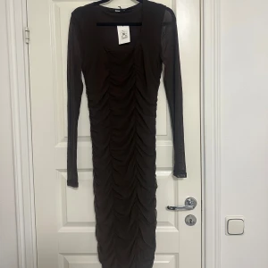 Brun långärmad klänning från Bikbok - Snygg brun klänning från Bikbok med lång ärm och rynkad design framtill. Perfekt för en stilren look. Klänningen har en kroppsnära passform och är tillverkad i ett lätt och bekvämt material.