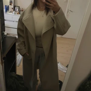 Beige kappa - Säljer en stilren beige kappa med långa ärmar och en klassisk krage. Perfekt för att hålla sig varm och snygg under kyligare dagar. Kappan har en elegant och tidlös design.