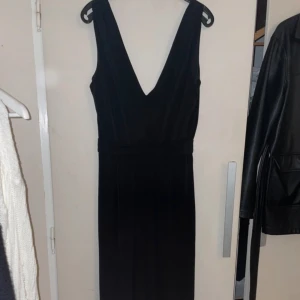 Svart byxdress  med v-ringning - Elegant svart byxdress med djup v-ringning och dragkedja i ryggen. Byxfressen har en stilren design och är perfekt för festliga tillfällen. Materialet ser ut att vara mjukt och följsamt.