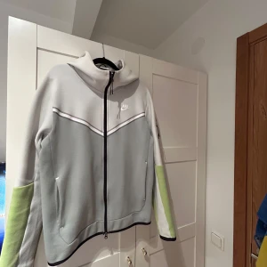 Grå och vit jacka från Nike - Säljer en stilren grå och vit jacka från Nike med dragkedja och huva. Jackan har långa ärmar med gröna detaljer och en liten Nike-logga på bröstet. Perfekt för en sportig look!