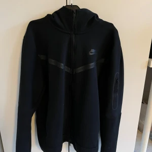 Nike tech fleece - Säljer en svart huvtröja från Nike med dragkedja och huva. Tröjan har långa ärmar och en diskret logga på bröstet. Tröjan är nästan helt oanvänd då den inte passar min stil. Köpt i butik och använd som sagt fåtal gånger. Hör av er om ni har frågor🌟👏