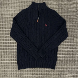 Ralph lauren half zip marin blå - Snygg svart stickad tröja från Ralph Lauren med kabelstickat mönster och en halv dragkedja framtill. Tröjan har långa ärmar och en liten röd logga på bröstet. Perfekt för våren, pris är ej hugget i sten och kom dm för frågor.