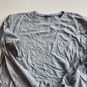 Grå långärmad tröja från Gant - Säljer en stilren grå långärmad tröja från Gant. Tröjan har en rund halsringning och ett diskret broderat Gant-emblem på bröstet. Perfekt för en avslappnad och snygg look.