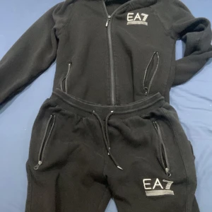 Svart hoodie och byxor från EA7 - Snygg svart hoodie med dragkedja och matchande byxor från EA7. Perfekt för en avslappnad stil. Hoodien har långa ärmar och byxorna har snörning i midjan för justerbar passform. Båda plaggen har EA7-loggan tryckt på framsidan.