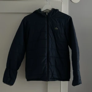 Svart quiltad jacka från Lacoste - Snygg svart quiltad jacka från Lacoste med dragkedja och huva. Perfekt för kyligare dagar med sin stilrena design och varma material. Jackan har långa ärmar och en diskret logga på bröstet.