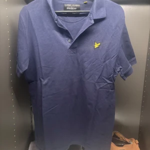 Blå pikétröja från Lyle & Scott - Säljer en stilren blå pikétröja från Lyle & Scott med korta ärmar och klassisk krage. Tröjan har en gul broderad logotyp på bröstet och knappar vid halsen. Perfekt för en avslappnad look.