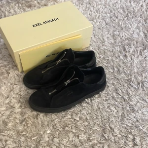 Svarta sneakers från Axel Arigato - Snygga svarta sneakers från Axel Arigato med dragkedja istället för traditionell snörning. Skorna har en stilren design med en solid svart sula och är tillverkade i skinn.