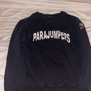 Svart tröja från Parajumpers - Säljer en svart tröja från Parajumpers med vit text på framsidan. Tröjan har långa ärmar och ett märke på ärmen. Perfekt för en stilren och avslappnad look.