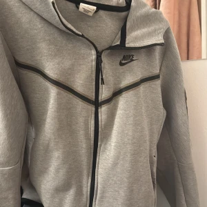 Grå hoodie från Nike - Säljer en grå hoodie från Nike med dragkedja och huva. Den har ett svart Nike-logotyp på bröstet och svarta detaljer längs dragkedjan och ärmarna. Perfekt för en avslappnad stil.
