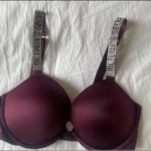 Vinröd/lilla bh från Victoria’s Secret med glittriga axelband - Snygg vinröd bh med vadderade kupor och glittriga axelband. Perfekt för att ge en touch av glamour till din outfit. Axelbanden är justerbara för bästa passform. Bilden är från när bh-n var ny så nu har den tappat några stenar men inget som är märkbart. Kan skicka bild på det vid intresse❤️