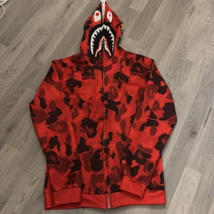 Röd camo hoodie med hajmotiv - Säljer en cool röd camo hoodie med hajmotiv på huvan. Hoodien har en dragkedja som går hela vägen upp och ett unikt mönster i rött och svart. Perfekt för dig som vill sticka ut! Tunnare material. 