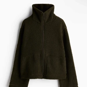 Olivgrön teddyjacka med dragkedja - Mysig olivgrön teddyjacka med dragkedja framtill. Jackan har en hög krage och långa ärmar, perfekt för kyligare dagar. Den har en avslappnad passform och stora fickor framtill för extra komfort och stil. Endast använd 2 gånger då den inte riktigt är min stil!