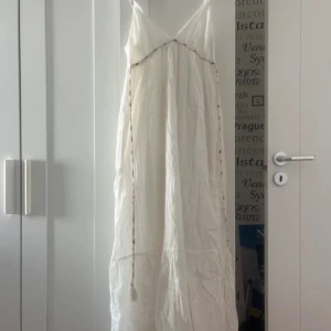 Vit långklänning med broderade detaljer - Vit långklänning med tunna axelband och broderade detaljer längs kanterna. 