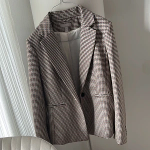 Rutig kavaj från H&M - Snygg rutig kavaj från H&M i beige, brun och svart. Kavajen har en klassisk passform med knappar framtill och vid ärmsluten. Perfekt för en stilren look.