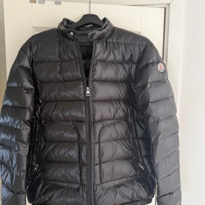 Svart dunjacka från Moncler - Säljer en stilren svart dunjacka från Moncler. Jackan har en quiltad design med dragkedja framtill och en hög krage. Perfekt för kyligare dagar med sin lätta och varma fyllning av vitt gåsdun. Köpt i andra hand men innan dess var den ny. Andvänd fåtal gånger bara! Skick är 9/10!