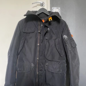 Svart jacka från Parajumpers - Säljer en svart vinterjacka från Parajumpers med huva och flera praktiska fickor. Jackan har dragkedja och knappar framtill samt en cool detalj med orange flärp vid kragen. Den har bara blivit använd av mig något fåtal gånger då jag inte hade någon användning av den.