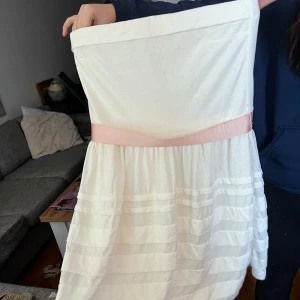 Vit axelbandslös klänning från Rut m.fl. - Säljer en söt vit axelbandslös klänning från Rut m.fl. med rosa band i midjan och dragkedja bak. Klänningen har ett fint randigt mönster på kjolen och är perfekt för sommarens festligheter. 🌸