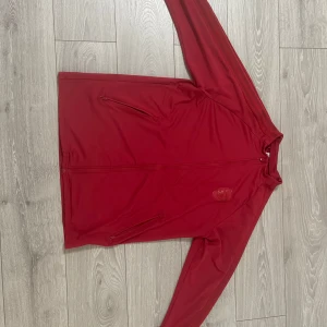 Röd kofta med dragkedja från adidas/arsenal - Snygg röd jacka med dragkedja framtill och två praktiska fickor. Perfekt för en sportig look eller en avslappnad dag ute. Jackan har långa ärmar och en stilren design.