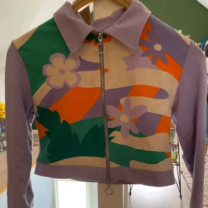 Färgglad croppad långärmad med blommönster - Säljer en unik och färgglad croppad långärmad med blommönster i lila, orange, grön och beige. Tröjan har en dragkedja framtill och långa ärmar. Står storlek S men fungerar på XS också