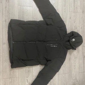 Svart dunjacka från Sail Racing - Säljer en svart dunjacka från Sail racing. Jackan har en dragkedja framtill och en justerbar huva. Perfekt för kalla dagar med sin varma och vadderade design. Den har långa ärmar och en stilren look.