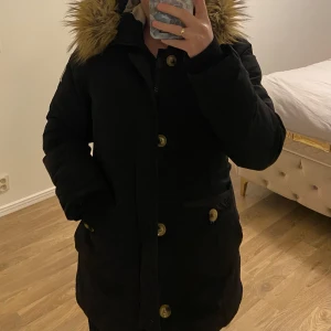 Svart parkajacka med fuskpäls - Snygg svea (MISS SMITH) svart parkajacka med fuskpäls på huvan och knappar framtill. Jackan har långa ärmar och praktiska fickor. Perfekt för kyliga dagar.