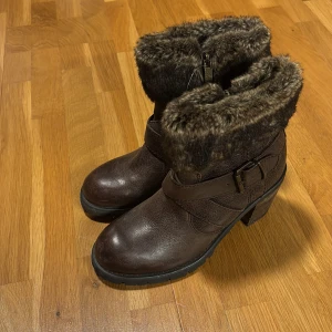 Bruna boots med pälsdetalj från 5th Avenue - Snygga bruna boots från 5th Avenue med pälsdetalj och dragkedja. De har en robust klack och ett dekorativt bälte runt skaftet. Perfekta för kyligare dagar med sin varma och stiliga design. Aldrig använda så är i väldigt bra skick.