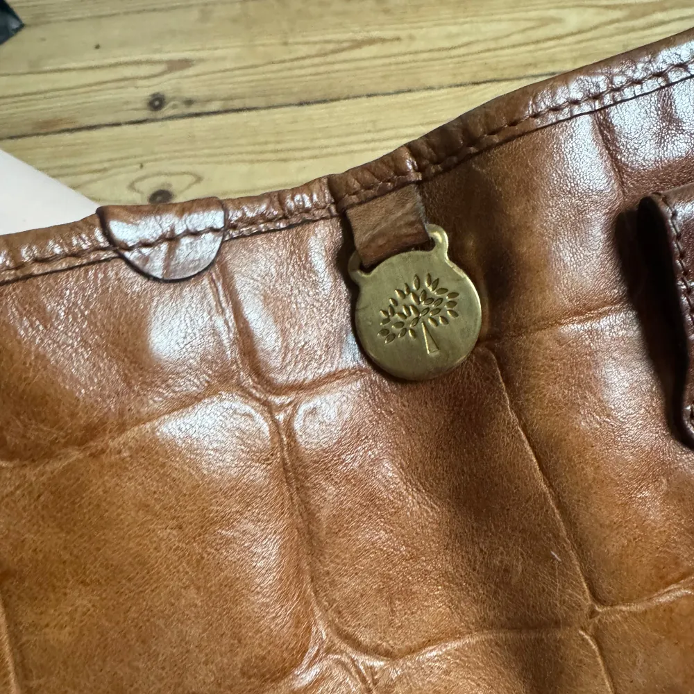 Snygg brun axelväska i skinn med krokodilpräglat mönster. Väskan har ett emblem på framsidan och en metallknapp med ett trädmotiv. Insidan är fodrad med ett rutigt tyg. Perfekt för en stilren look. ÄKTA. . Laukut & Käsilaukut.