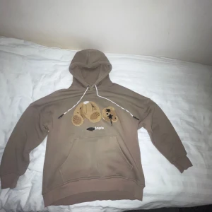 Beige hoodie från Palm Angels - Säljer en beige hoodie från Palm Angels med en unik nallebjörnsdesign på framsidan. Hoodien har en klassisk känguruficka och justerbar huva med vita snören. Perfekt för en avslappnad stil. Går och sänka priset vid snabb affär.