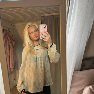 Beige prickig blus - Säljer en söt beige blus med små prickar och svarta detaljer. Blusen har långa ärmar och en lös passform, perfekt för en avslappnad stil. Den är lätt och luftig, vilket gör den idealisk för vår och sommar.