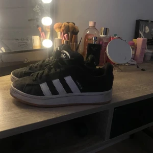 Svarta Adidas sneakers med vita ränder - Säljer ett par klassiska svarta Adidas sneakers med vita ränder och brun sula. Skorna har snörning och den ikoniska Adidas-loggan på hälen. Perfekta för en avslappnad stil. MÖTS UPP I TYRESÖ STOCKHOLM, de tvättas såklart innan köp