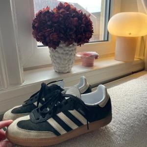 Adidas Samba sneakers - Snygga Adidas Samba sneakers i svart och beige med klassiska tre ränder i vitt. Skorna har snörning och en gummisula som ger bra grepp. Perfekta för en stilren och sportig look.
