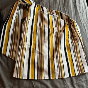 Randig oneshoulder topp - Snygg oneshoulder topp med randigt mönster i svart, vitt, gult och brunt. Perfekt för en trendig look med sin unika design och färgkombination.