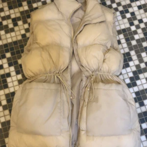 Beige dunväst från McKinley - Säljer en beige dunväst från McKinley med dragkedja och knytning i midjan. Perfekt för kyligare dagar och ger en stilren look. Västen är vadderad och håller dig varm.