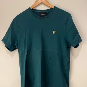 Grön t-shirt från Lyle & Scott - Snygg grön t-shirt från Lyle & Scott med ett gult broderat emblem på bröstet. Använd ett par gånger men är i bra skick och inget dåligt. Sälja för ett billigt pris så var snabb med att köpa! 